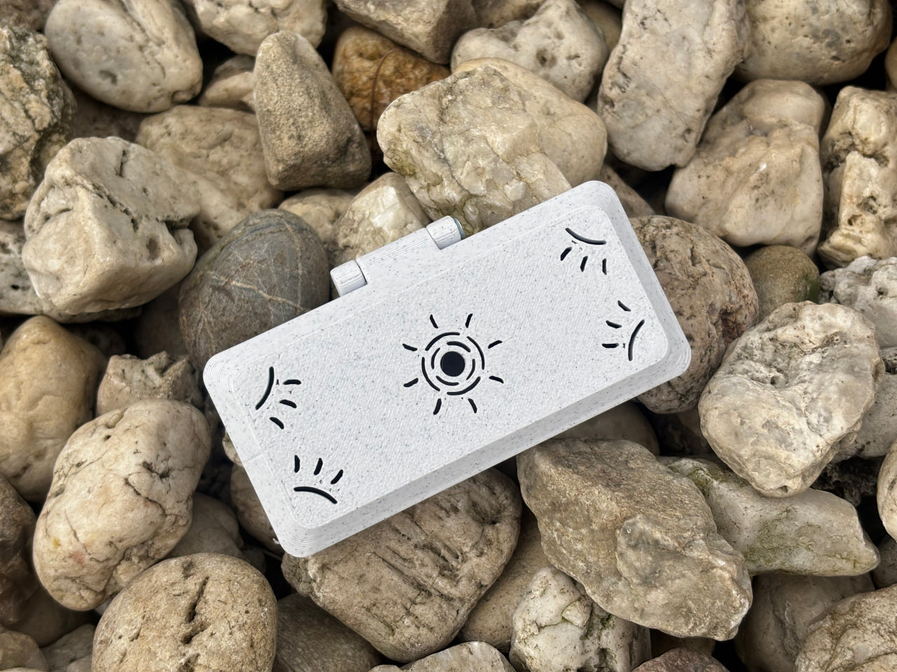 The Reed Case for 3 Legere Oboe Reeds by FswStudio, featuring white rectangular handmade design with sun and curved line motifs, is displayed on a bed of light-colored, irregularly shaped rocks.