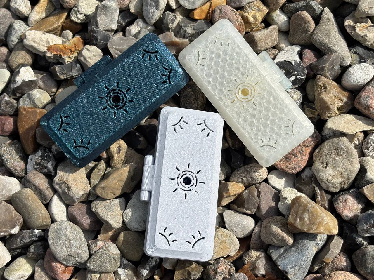Three patterned, rectangular Reed Case for 3 Oboe Reeds by FswStudio—dark green, off-white, and transparent—rest on colorful rocks. Each features eye motifs and a Sun series-inspired circular design at the center.