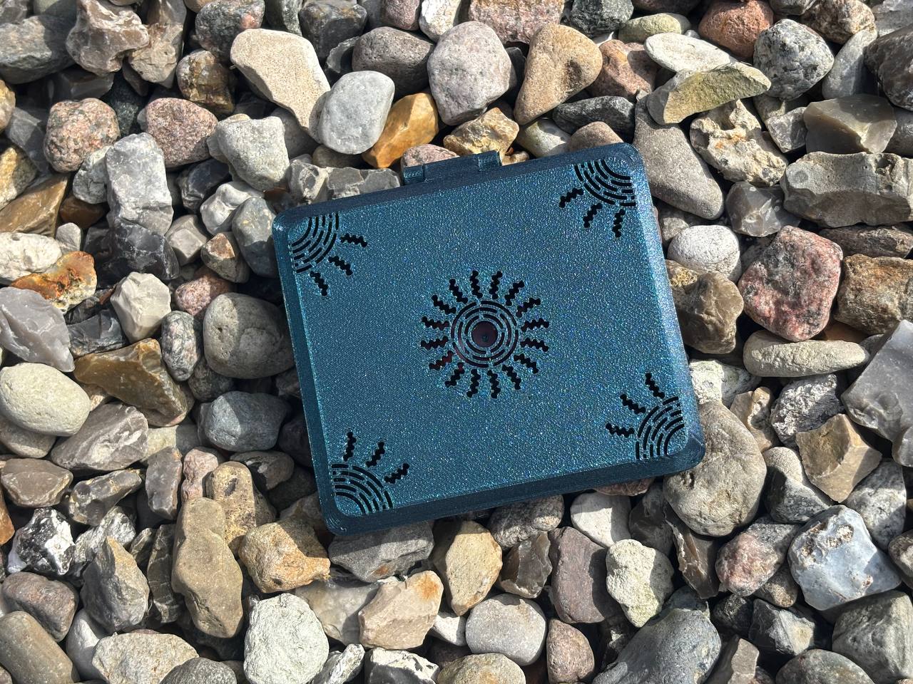 The FswStudio Reed Case for 10 Oboe Reeds from the Sun series features dark teal, rectangular handmade design with black sun motifs, showcased on multicolored rocks as sunlight highlights its unique texture.