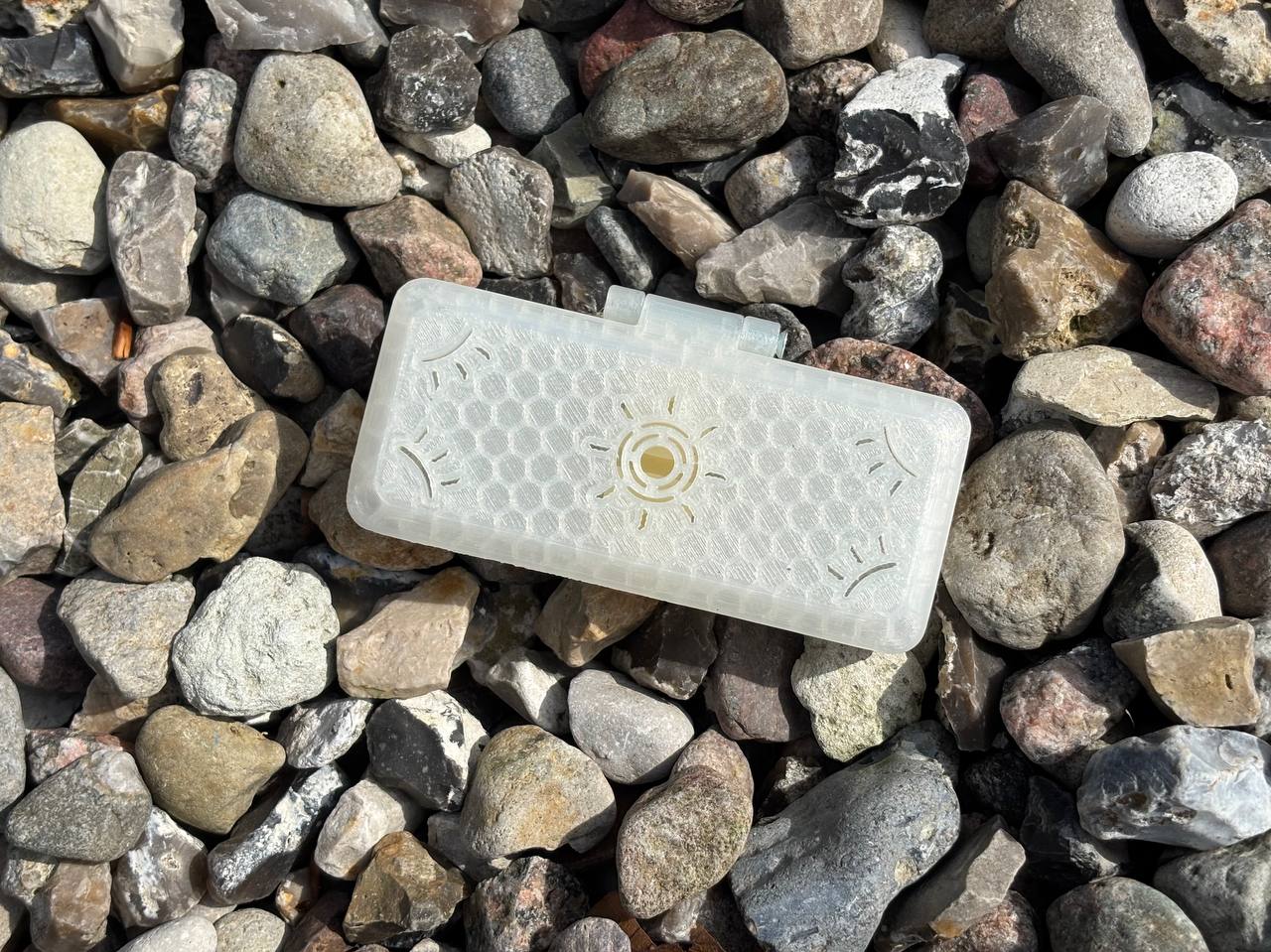 The Reed Case for 3 Oboe Reeds by FswStudio features a translucent, handmade design with a honeycomb pattern and circular emblem, displayed on multicolored textured stones.