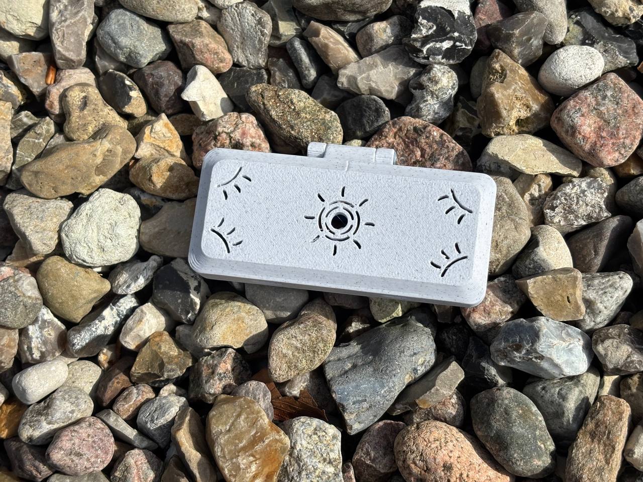 A handmade rectangular gray plastic Reed Case for 3 Oboe Reeds by FswStudio, featuring sun-like patterns from the Sun series, rests on colorful rocks in bright sunlight.