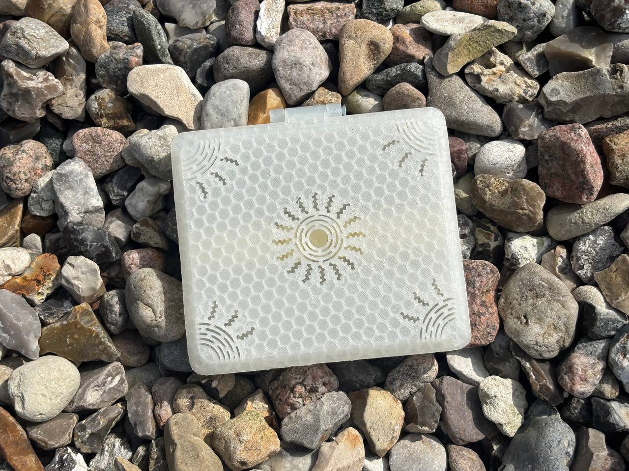 The FswStudio Reed Case for 10 Oboe Reeds, from the Sun series, is a translucent white square with a honeycomb pattern and gold sun designs, shown resting on assorted rocks—ideal for storing handmade oboe reeds.