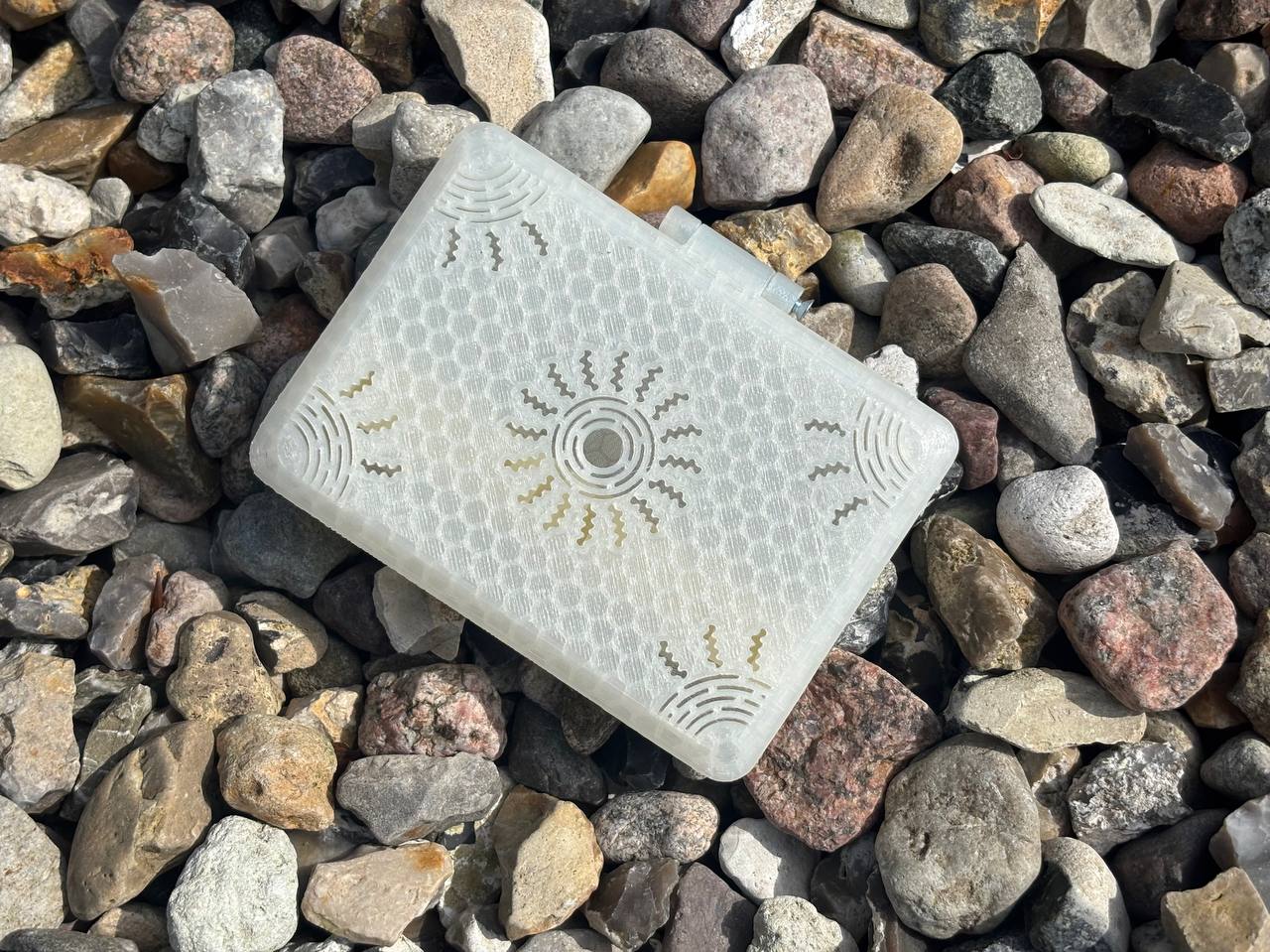 A rectangular, textured white FswStudio Reed Case for 10 English Horn Reeds from the Sun series, carved with sun and wave symbols, rests on colorful rocks as sunlight casts shadows and highlights its detailed patterns.