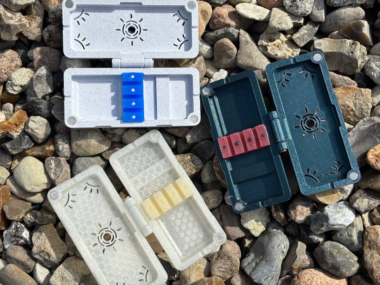 Three rectangular Reed Cases for 3 Oboe Reeds by FswStudio—gray, dark green, and translucent white—are open and arranged on small rocks. Each case holds three colorful oboe reeds.