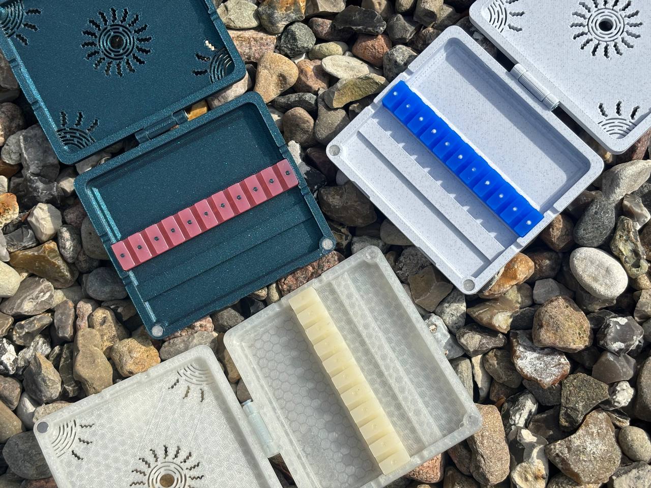 Three open FswStudio Reed Case for 10 Oboe Reeds, each with decorative vented lids, display colorful pink, blue, and white oboe reeds arranged on rocky ground.