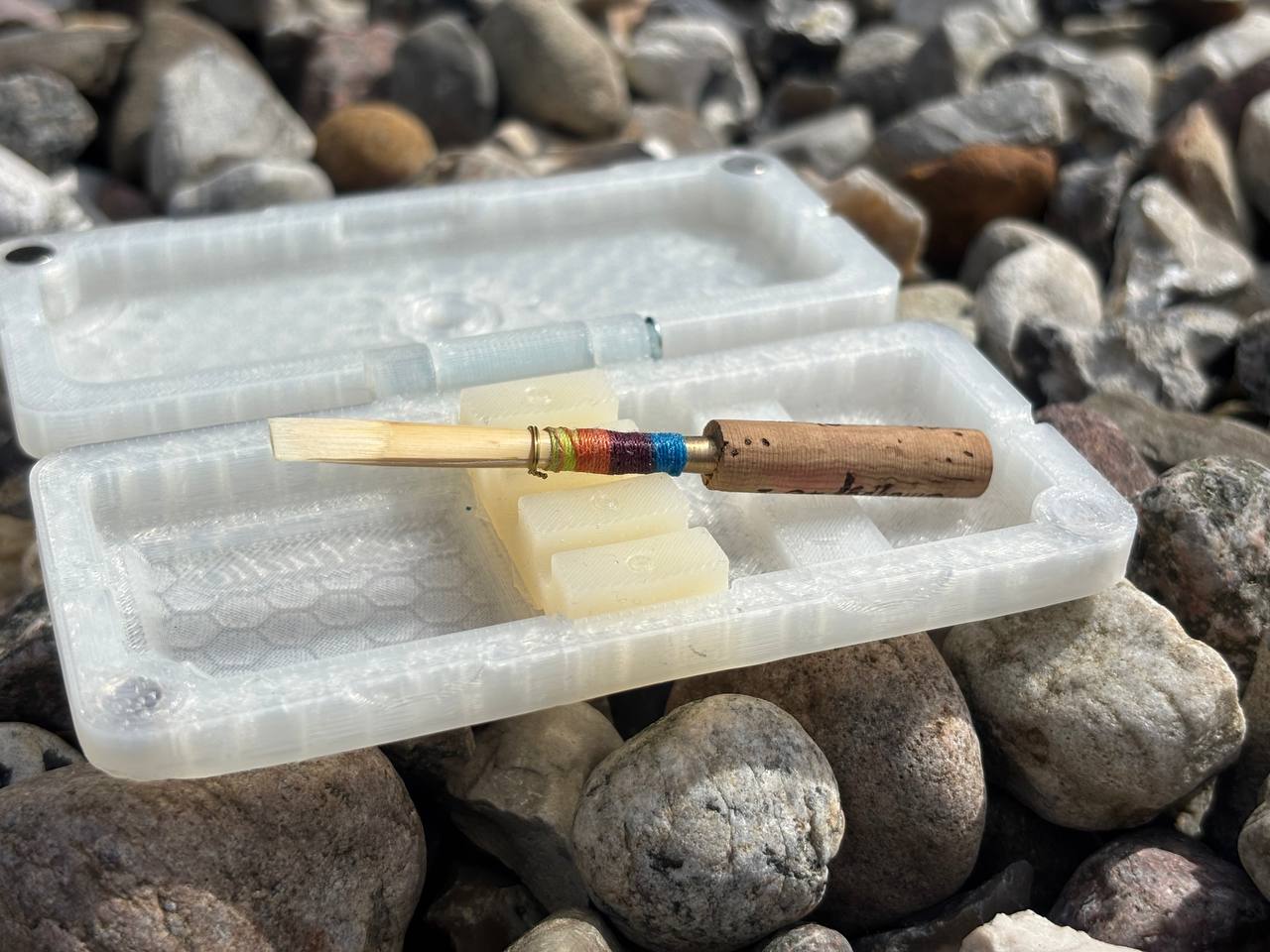 A handmade oboe reed sits on an open FswStudio Reed Case for 3 Oboe Reeds, featuring a white honeycomb pattern, surrounded by small rocks in bright sunlight.