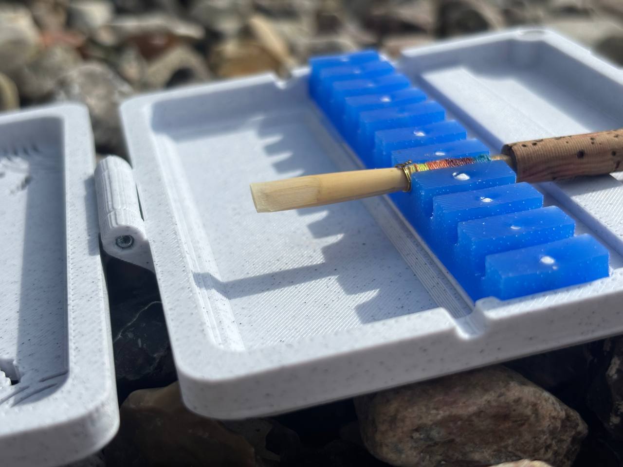 A close-up of an oboe reed drying on a blue holder inside a white plastic FswStudio Reed Case for 10 Oboe Reeds, with pebbles scattered in the background.