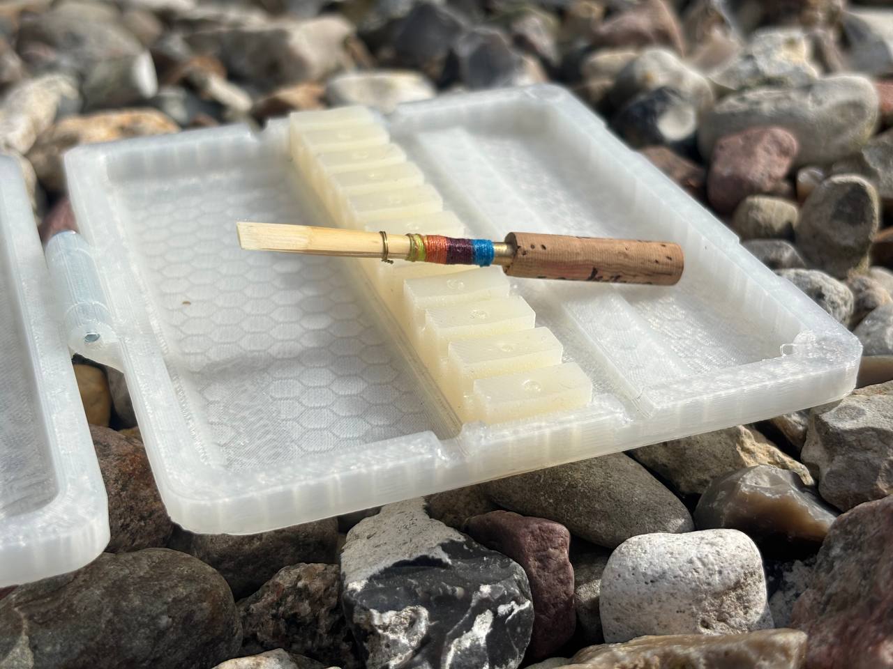 A FswStudio Reed Case for 10 Oboe Reeds rests on a surface covered with assorted rocks, its translucent plastic design contrasting the colorful scenery.