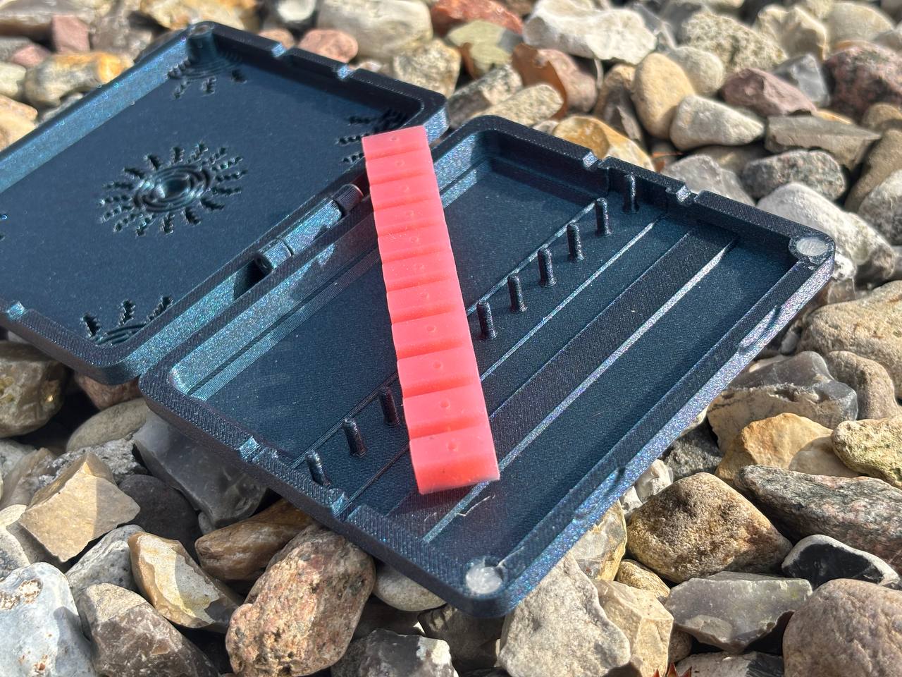 A black handmade FswStudio Sun Reed Case for 10 Oboe Reeds with an open lid lies on rocks, holding a red insert with small dots and vertical pegs for oboe reeds inside.