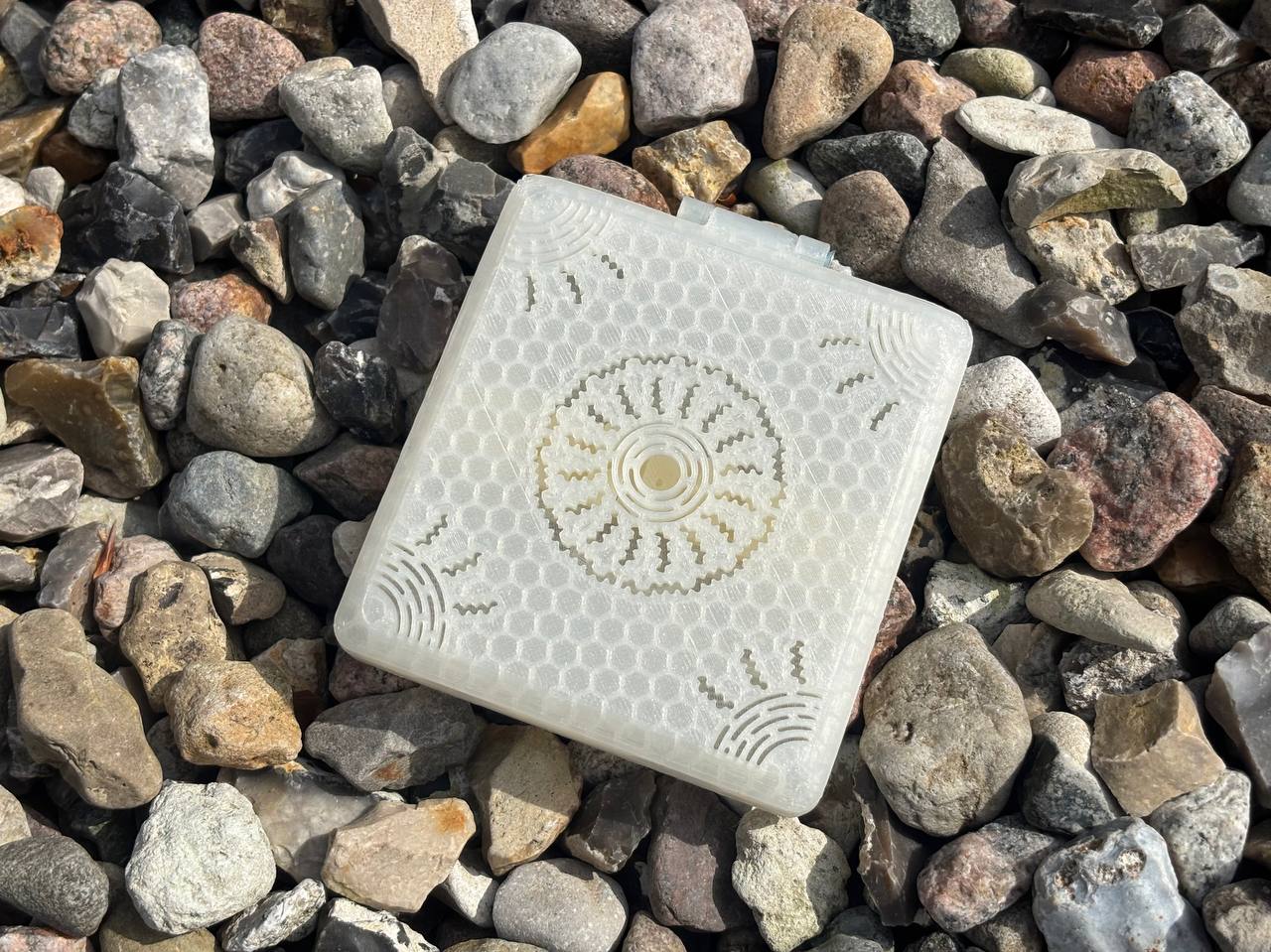 The FswStudio Combined Reed Case for 5 Oboe and 3 English Horn Reeds, a square translucent accessory with a geometric design, rests on gray and brown stones, its pattern catching the light.