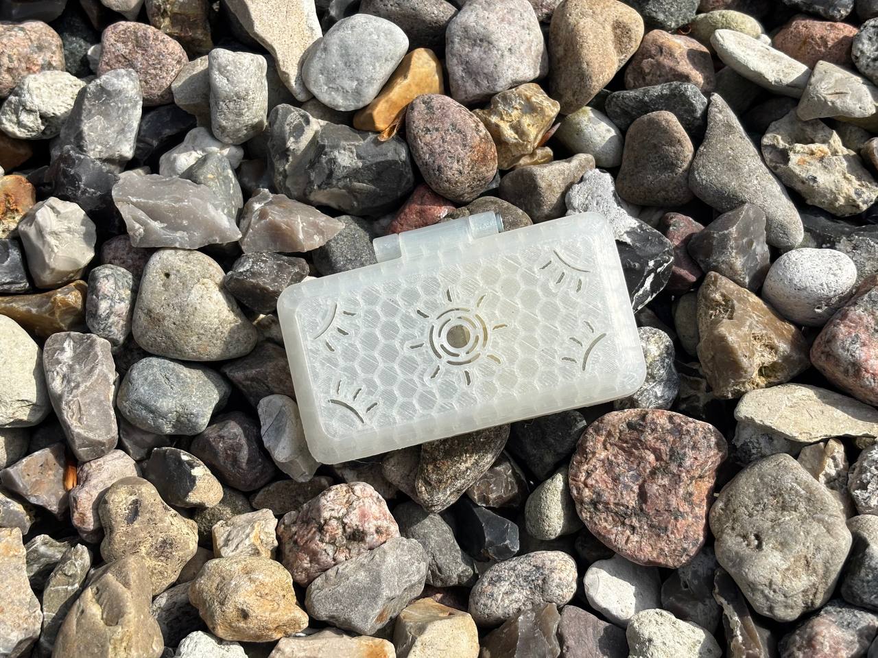 A translucent, rectangular FswStudio Reed Case for 3 English Horn Reeds with a honeycomb pattern and circular vent rests on colorful rocks. Sunlight accents its texture and that of the stones—perfect storage for your English Horn reeds.