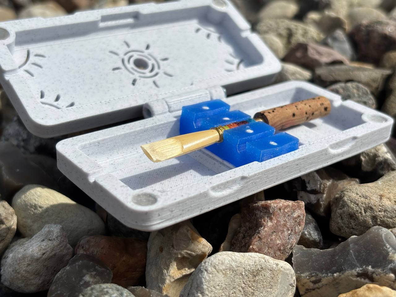 An FswStudio Reed Case for 3 Oboe Reeds holds a single wooden-base oboe reed in a blue holder, displayed open on assorted outdoor rocks.