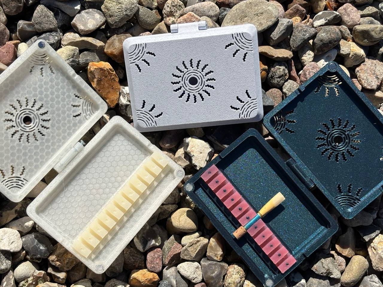 Three small FswStudio vented plastic Reed Cases for 10 English Horn Reeds display geometric patterns on rocks. Two cases hold harmonica reeds—one with light, one red-brown reeds and a tool—while the third Sun series box is empty.