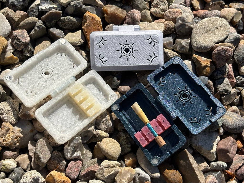 Three small, decorative FswStudio Sun series Reed Cases for 3 English Horn Reeds sit open on gravel. One handmade case holds a rolled item, one contains three English Horn reeds, and the third is empty. Sunlight casts shadows over the scene.