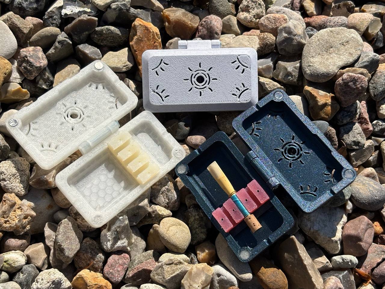 Three small, decorative FswStudio Sun series Reed Cases for 3 English Horn Reeds sit open on gravel. One handmade case holds a rolled item, one contains three English Horn reeds, and the third is empty. Sunlight casts shadows over the scene.