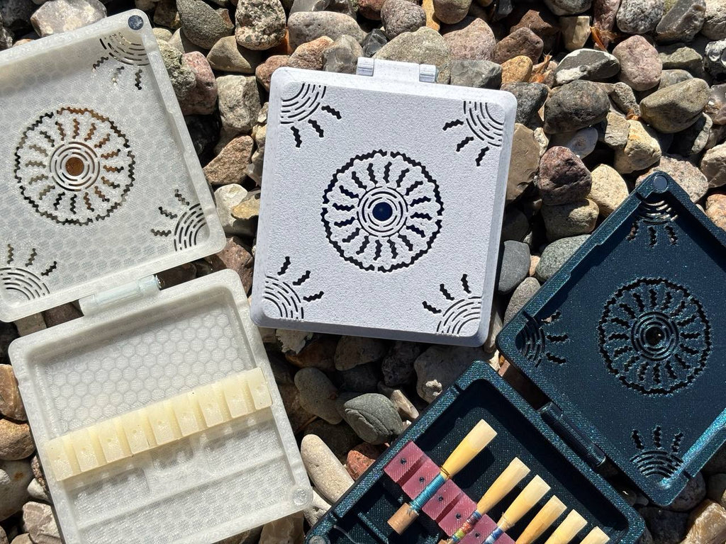 FswStudio’s Combined Reed Case for 5 Oboe and 3 English Horn Reeds features intricate circular designs. Resting on rocks in sunlight, two cases are open to display reeds inside while the others remain closed, highlighting their artisan craftsmanship.
