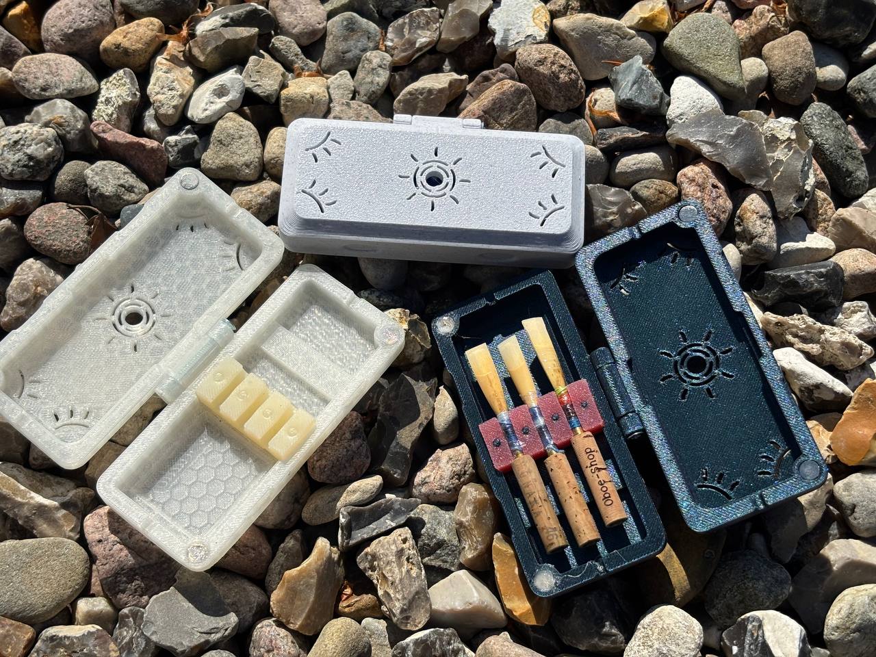 Three handmade FswStudio Reed Case for 3 Oboe Reeds from the Sun series (white, gray, black) are displayed open on gravel. The black case holds four oboe reeds; the others are empty or hold reed-shaped blocks.