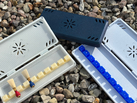 Three rectangular FswStudio Reed Cases for 9 contrabassoon reeds rest on small stones. Two open cases display reeds and extra slots, while the closed dark blue case features a handmade flower-shaped cutout design.
