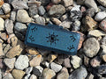 The FswStudio Reed Case for 3 Oboe Reeds is a small, dark green handmade box featuring eye and Sun designs on its lid, shown on multicolored stones—ideal for safely storing your oboe reeds.