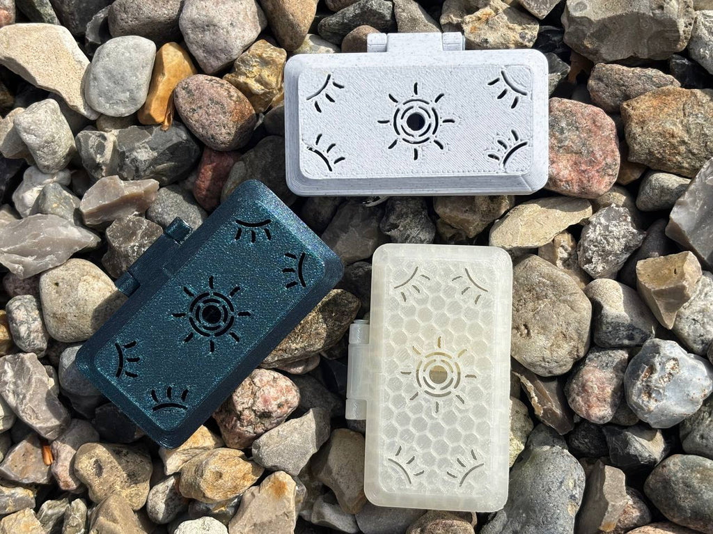 Three FswStudio Reed Cases for 3 English Horn Reeds are shown on gray and brown rocks. Each Sun series case is small, rectangular, and plastic—available in black, white, or translucent with unique eye and sun designs.