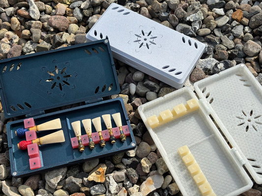 Two open handmade FswStudio Combined Reed Cases for 5 bassoon and 3 contrabassoon reeds rest on gravel, displaying assorted colored reeds. A closed case is nearby.