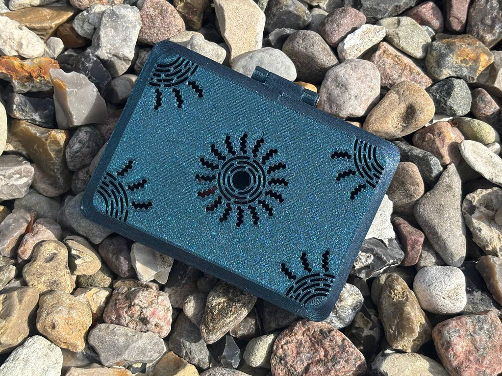 A small black FswStudio Reed Case for 10 English Horn Reeds, handmade with sun and corner designs, sits slightly askew on colorful rocks. Its sparkly Sun series motif catches the light while the box remains closed among the stones.