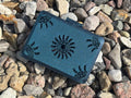 A small black FswStudio Reed Case for 10 English Horn Reeds, handmade with sun and corner designs, sits slightly askew on colorful rocks. Its sparkly Sun series motif catches the light while the box remains closed among the stones.