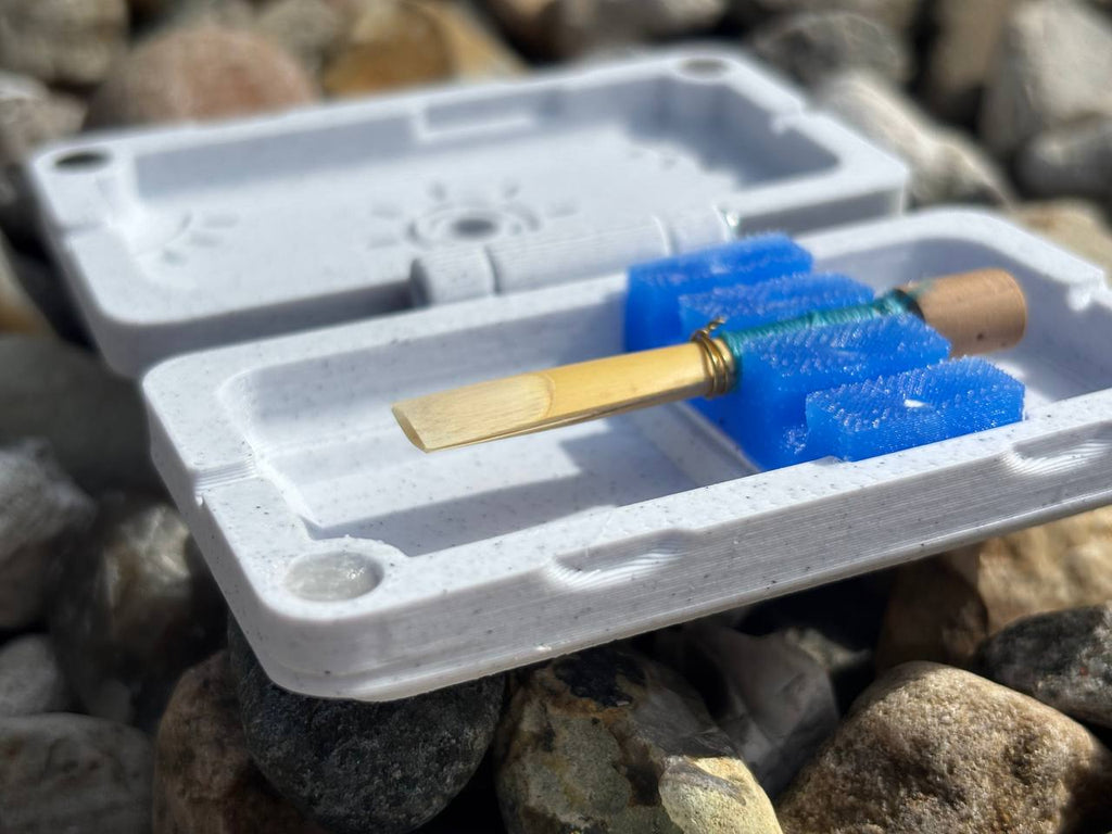 A close-up of the FswStudio Reed Case for 3 English Horn Reeds from the Sun series sits on rocks, sunlight casting shadows over its white plastic shell and blue foam insert, emphasizing each objects distinct textures.