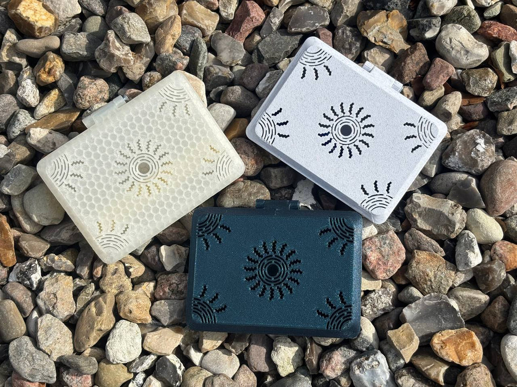 Three rectangular FswStudio Reed Cases for 10 English Horn Reeds from the Sun series, featuring abstract sunburst designs in white honeycomb, light gray, and dark green, rest on a bed of small rocks.