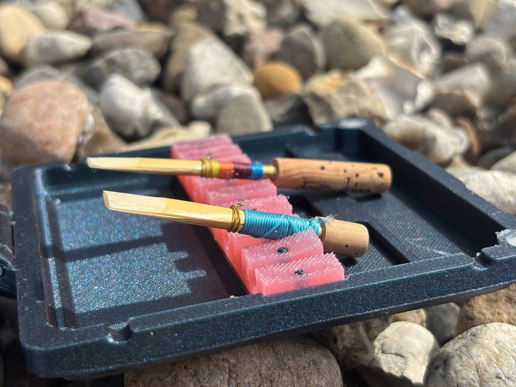 Two oboe reeds with wrapped handles rest on an FswStudio Combined Reed Case for 5 Oboe and 3 English Horn Reeds, handmade and lined with pink foam, set atop rocks of varying sizes and colors.