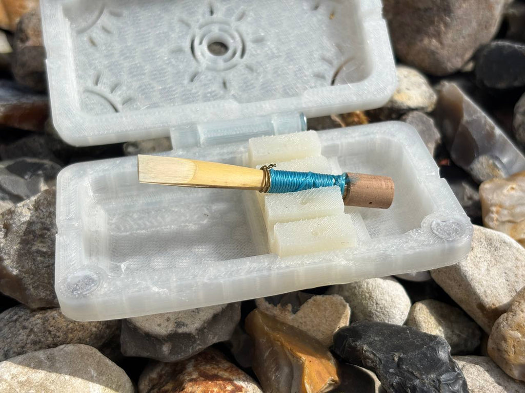 A FswStudio Reed Case for 3 English Horn Reeds rests open on rocks in sunlight, holding double reeds with blue thread at the base.