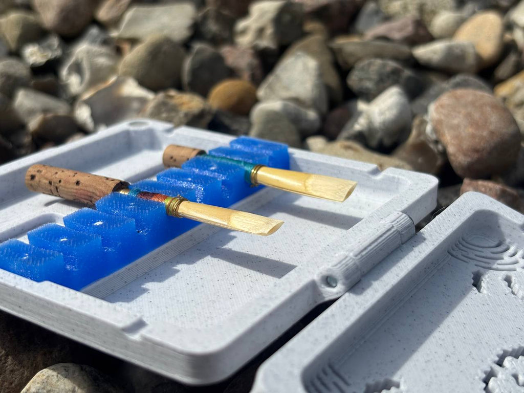 The FswStudio Combined Reed Case holds 5 oboe and 3 English horn reeds with cork bases in a blue plastic holder, shown outdoors on multi-colored rocks.