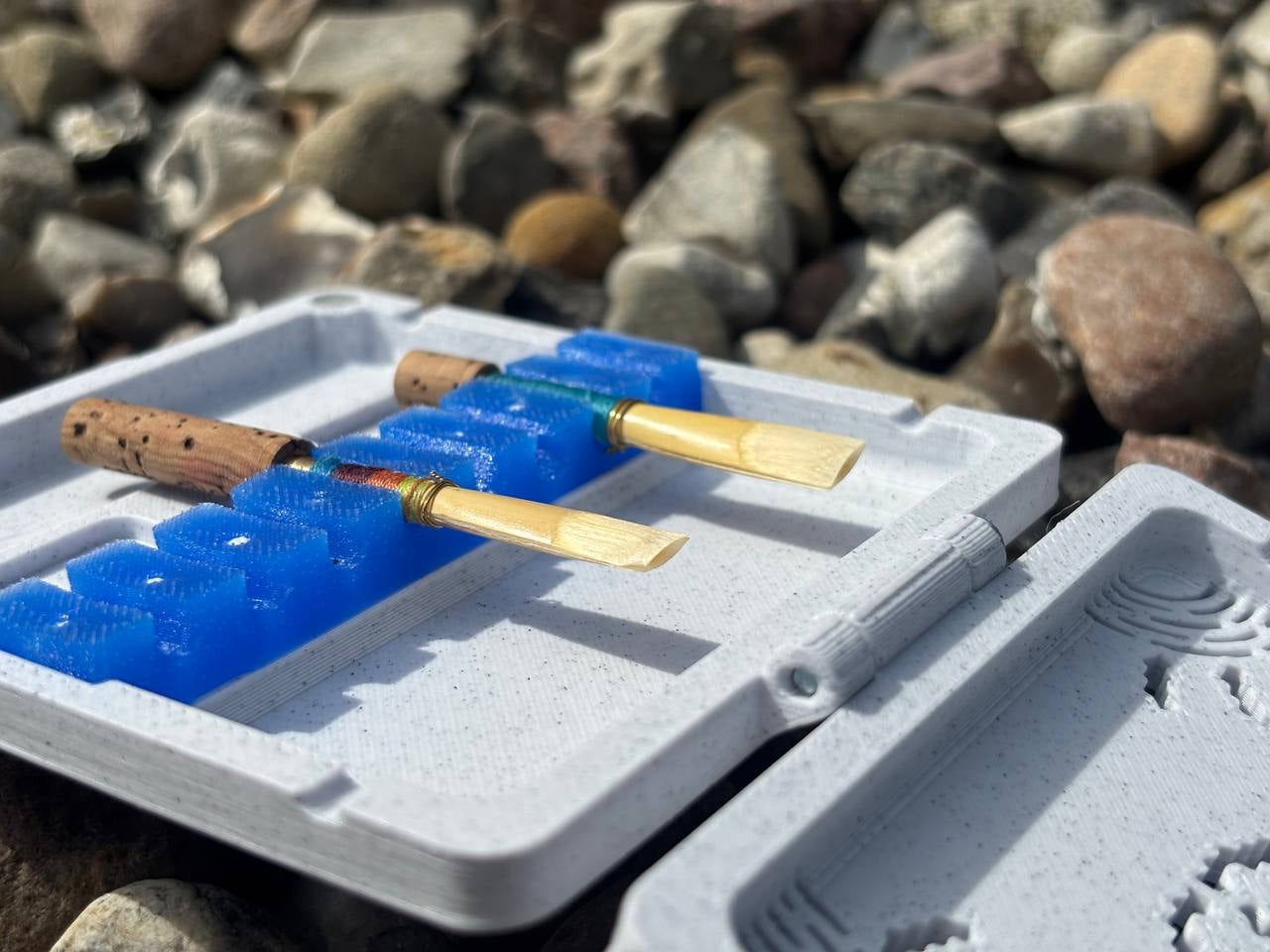 The FswStudio Combined Reed Case holds 5 oboe and 3 English horn reeds with cork bases in a blue plastic holder, shown outdoors on multi-colored rocks.