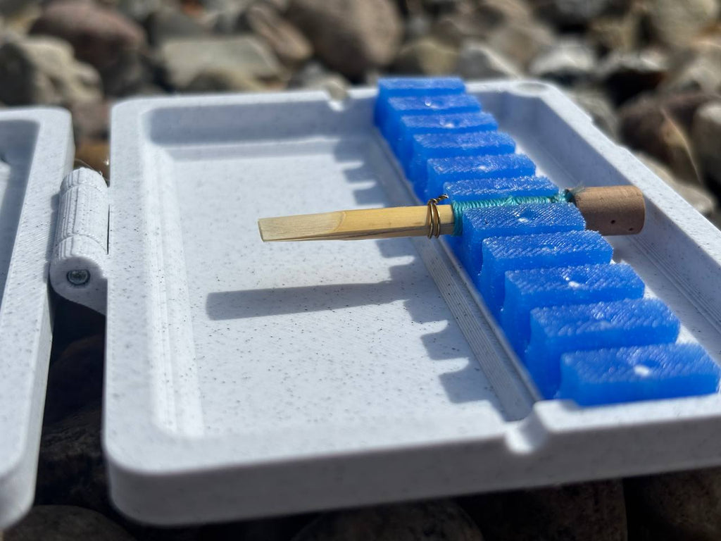 A close-up of a single English horn reed in a blue foam holder inside the FswStudio Reed Case for 10 English Horn Reeds, with rocks blurred in the background.