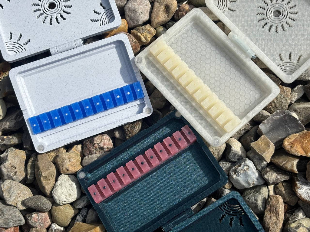 Three FswStudio Reed Cases for 10 English Horn Reeds, each with a patterned lid and compartments holding blue, white, and pink reeds, are displayed outdoors on small rocks.