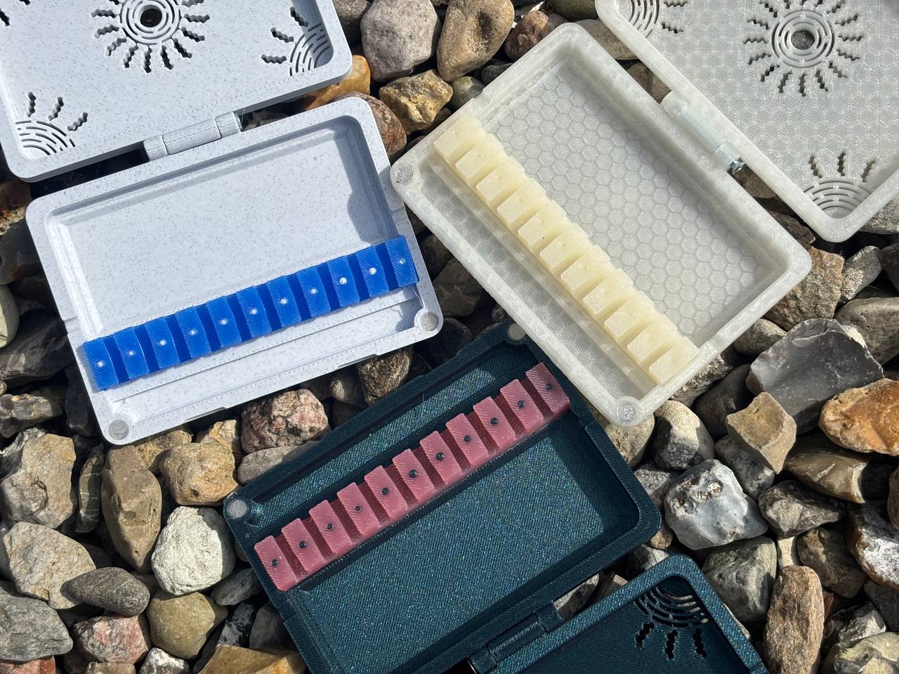 Three FswStudio Reed Cases for 10 English Horn Reeds, each with a patterned lid and compartments holding blue, white, and pink reeds, are displayed outdoors on small rocks.