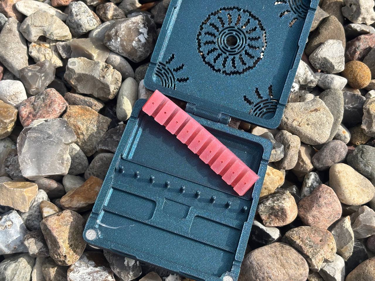 A FswStudio Combined Reed Case for 5 oboe and 3 English horn reeds, featuring a blue plastic handmade design with a decorative pattern, is open to show red reeds inside and rests on a surface with assorted rocks.