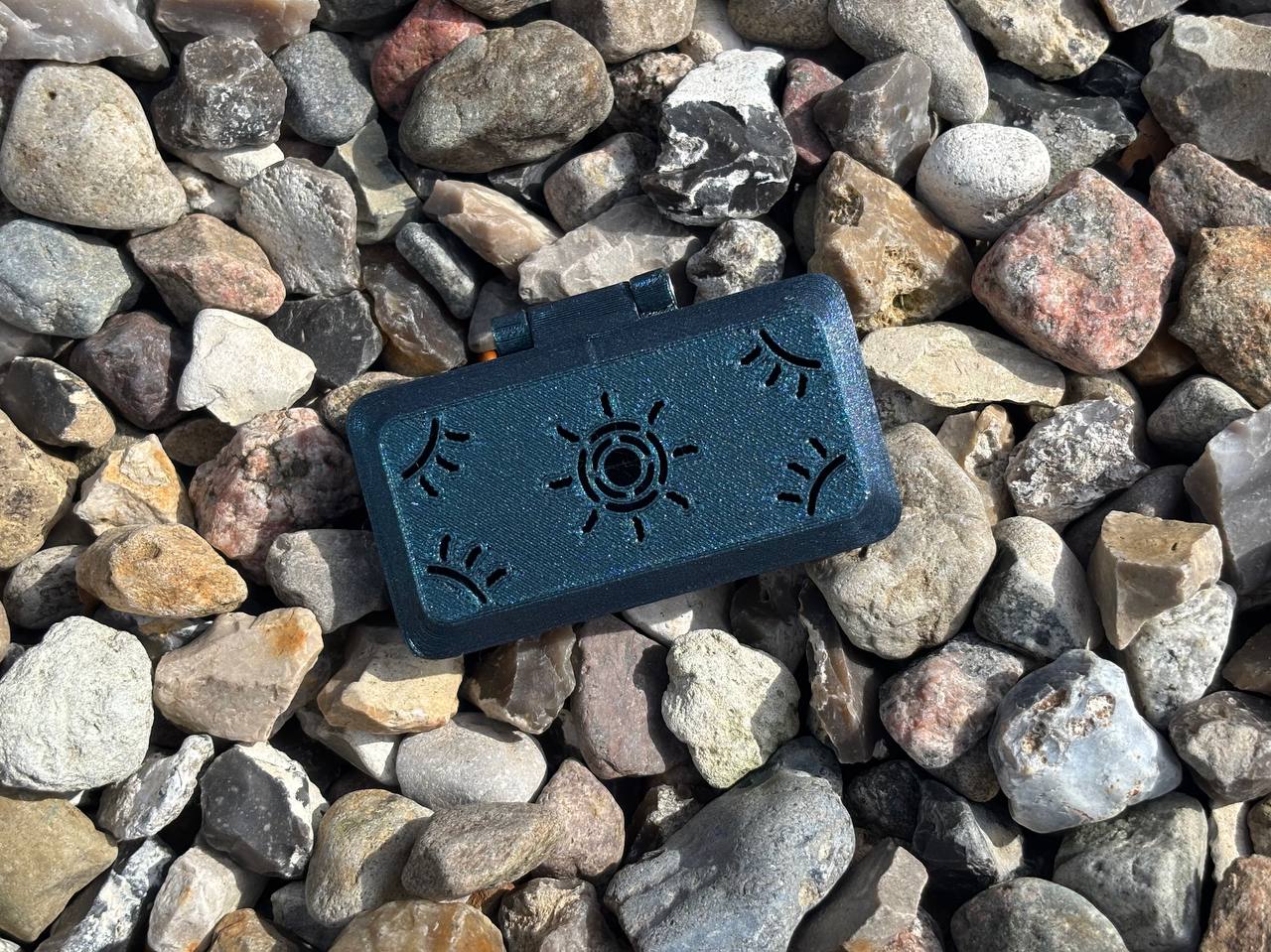 A small black Reed Case for 3 English Horn Reeds by FswStudio from the Sun series, featuring a sun and rays design, rests on multicolored rocks as sunlight casts shadows over the box and stones.
