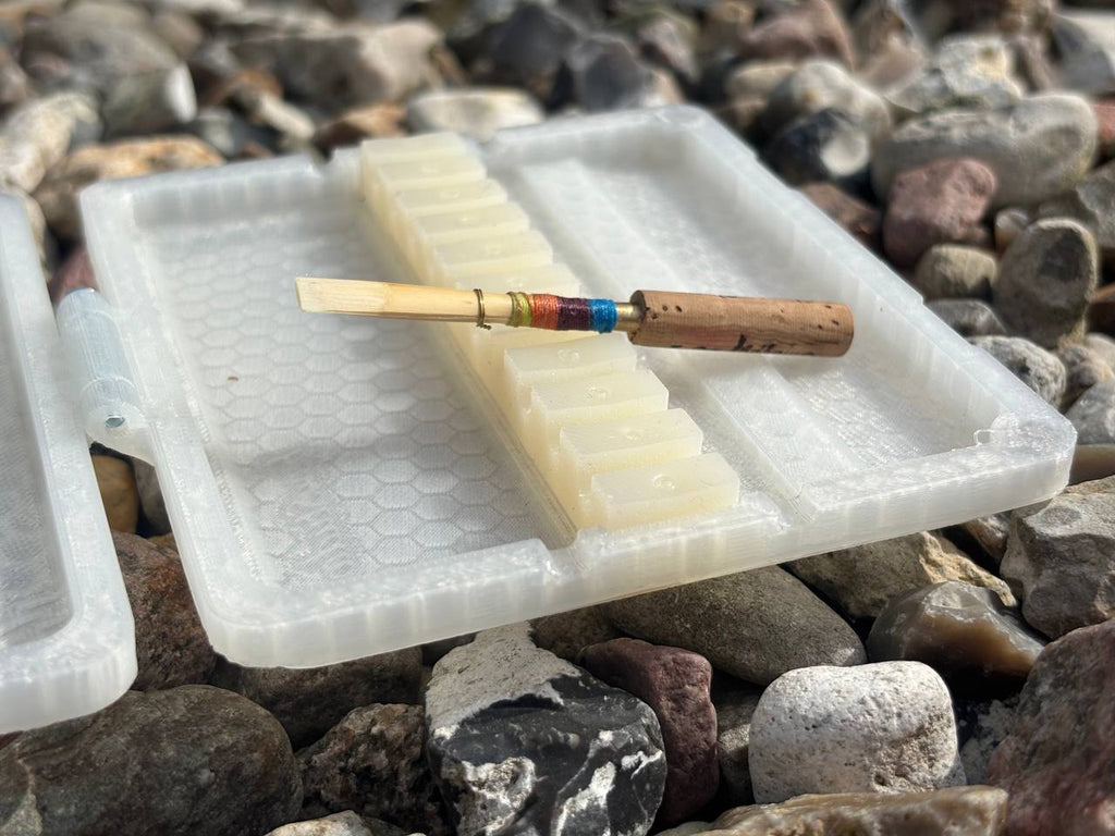 A FswStudio Reed Case for 10 Oboe Reeds rests on a surface covered with assorted rocks, its translucent plastic design contrasting the colorful scenery.