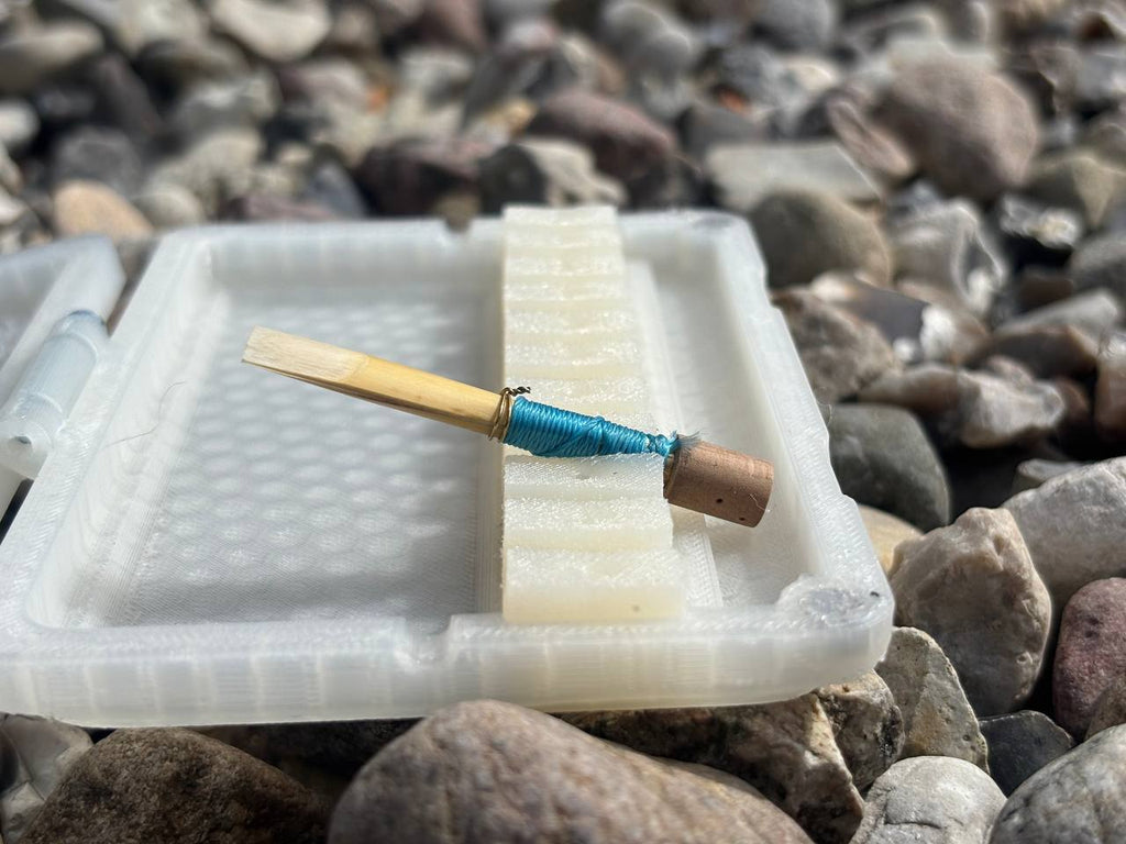 A close-up of a double reed from the Sun series with blue thread rests on an FswStudio Reed Case for 10 English Horn Reeds, surrounded by gray and brown pebbles.