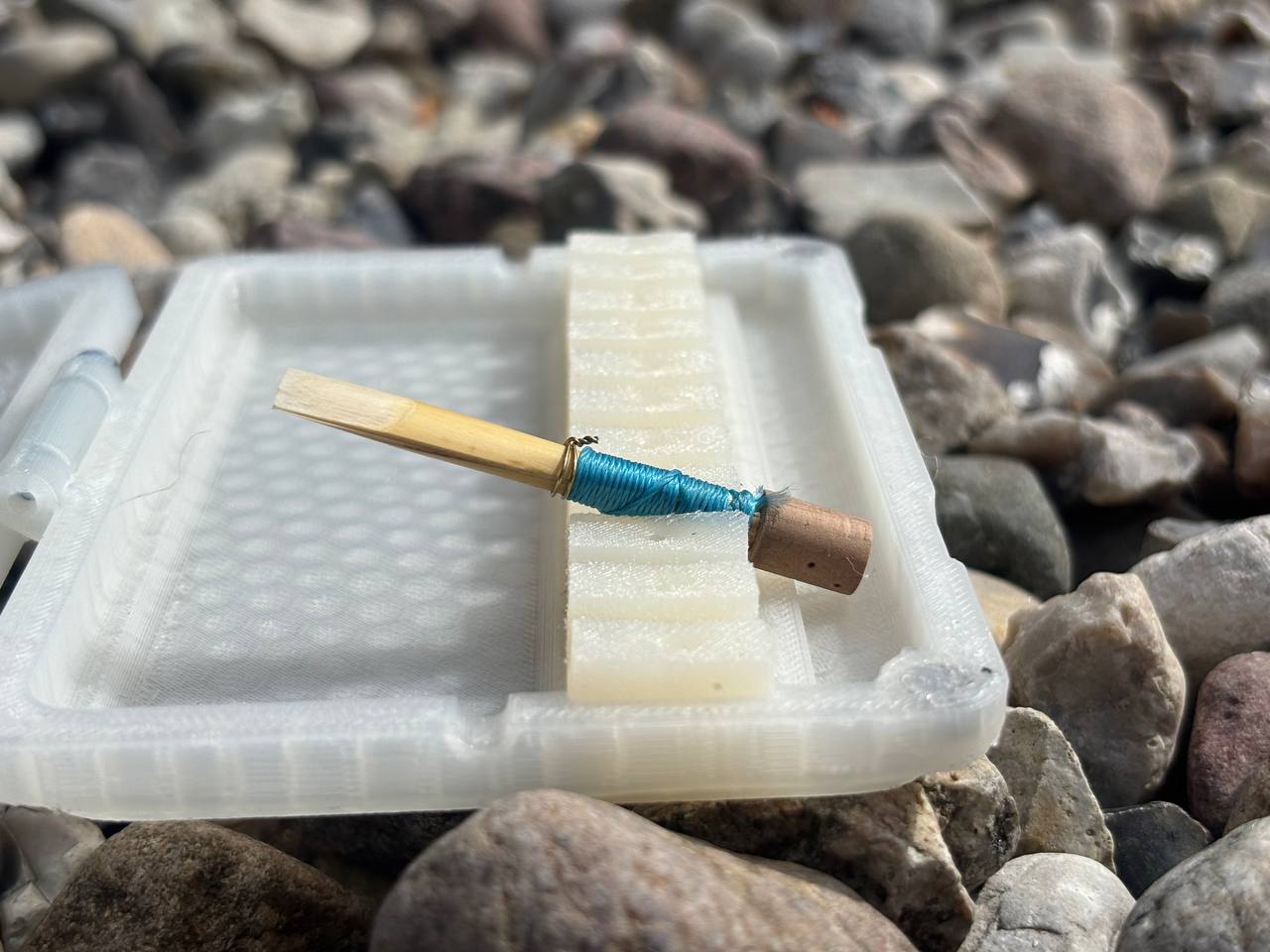 A close-up of a double reed from the Sun series with blue thread rests on an FswStudio Reed Case for 10 English Horn Reeds, surrounded by gray and brown pebbles.