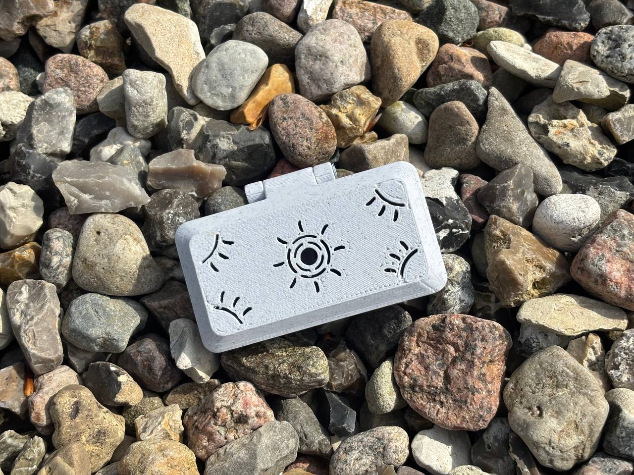The FswStudio Reed Case for 3 English Horn Reeds features handmade sun and eye designs on a white rectangular case, shown resting on multicolored rocks.