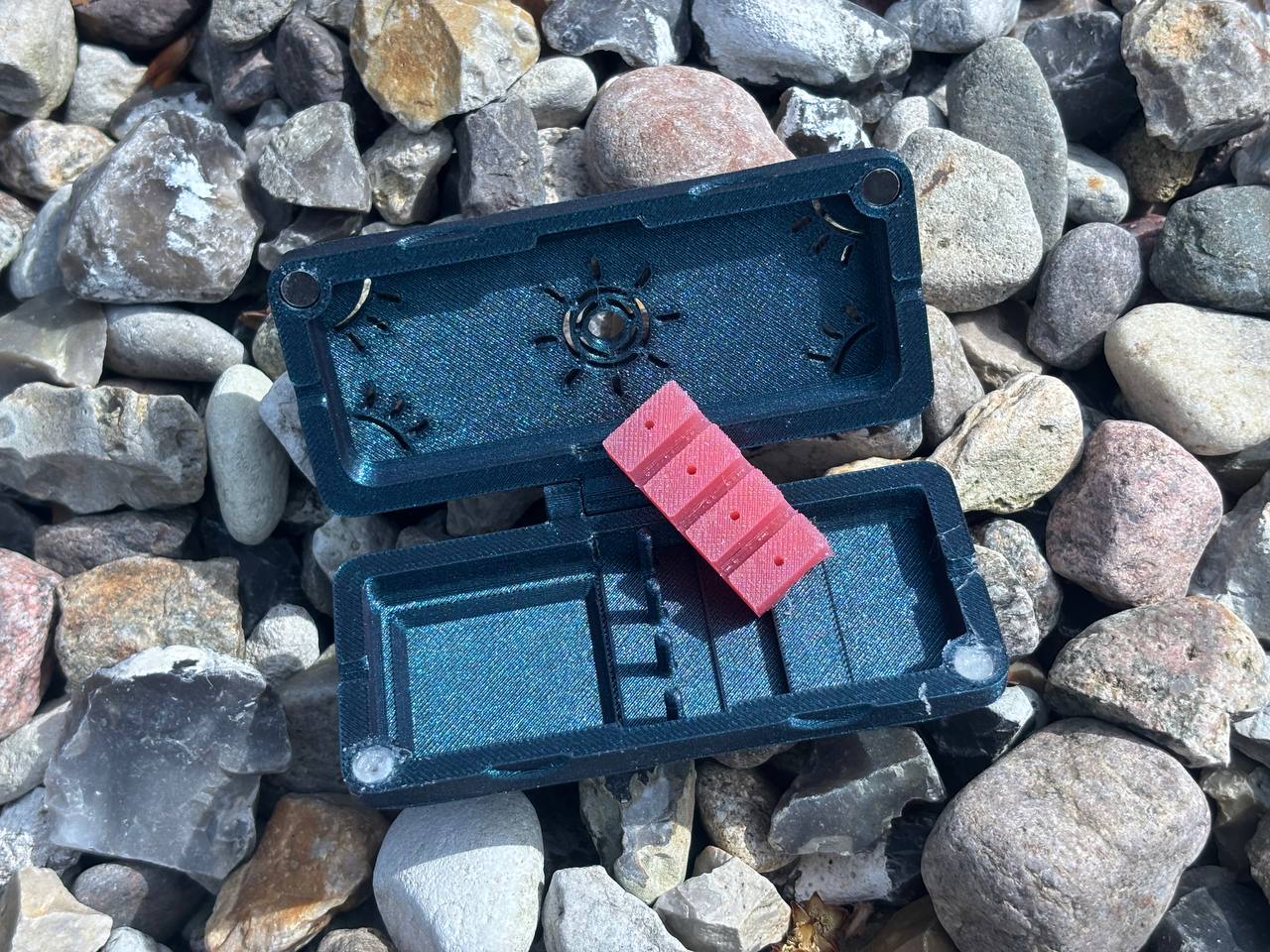 A compact, open Reed Case for 3 Oboe Reeds by FswStudio rests on a surface covered with colorful rocks and pebbles, displaying a vibrant red domino inside.