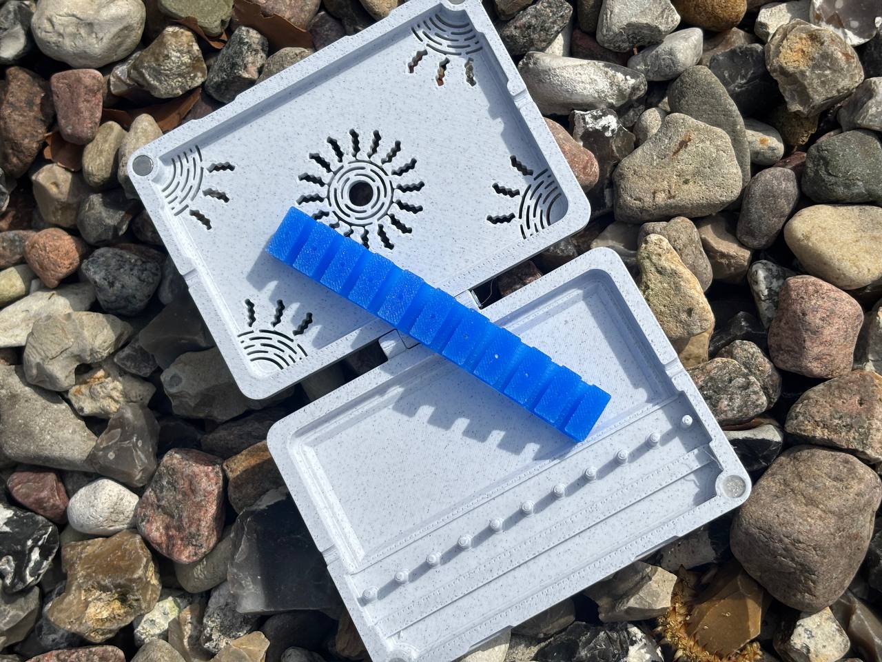 FswStudio’s gray plastic Reed Case for 10 English Horn Reeds, handmade and featuring sunburst vent patterns, lies open on rocks with a blue segmented strip inside—ideal for securely storing your reeds.