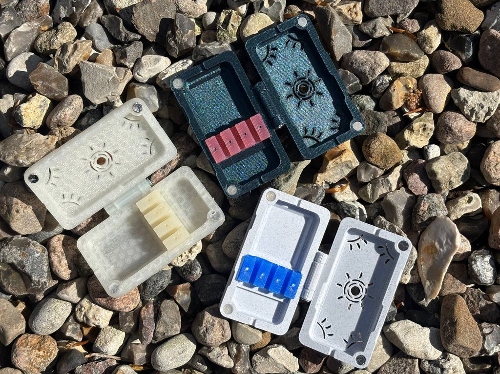 Three handmade FswStudio Reed Cases for 3 English Horn Reeds, each with a hinged lid and colored slots, are open on multicolored stones under sunlight.