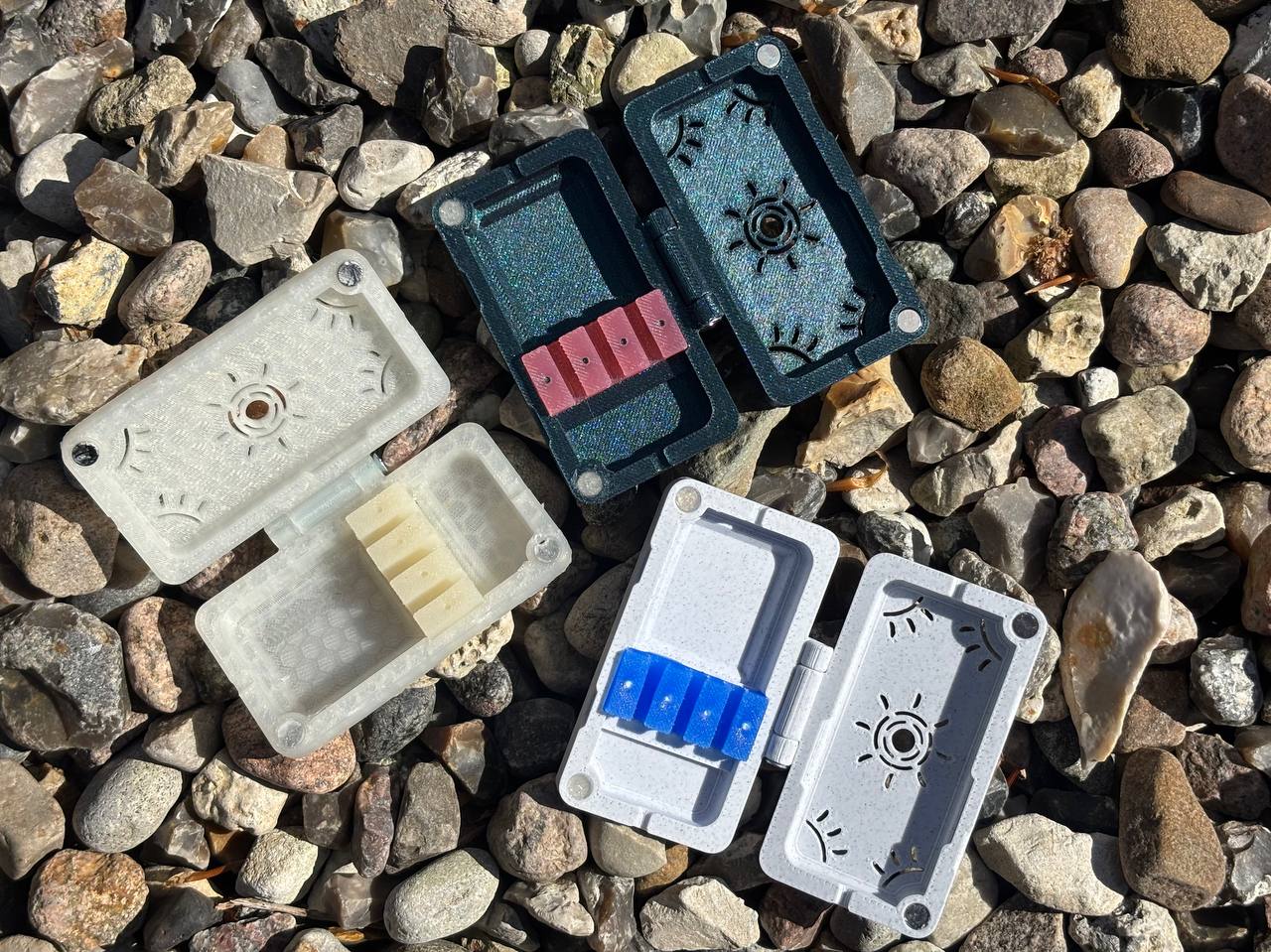 Three handmade FswStudio Reed Cases for 3 English Horn Reeds, each with a hinged lid and colored slots, are open on multicolored stones under sunlight.